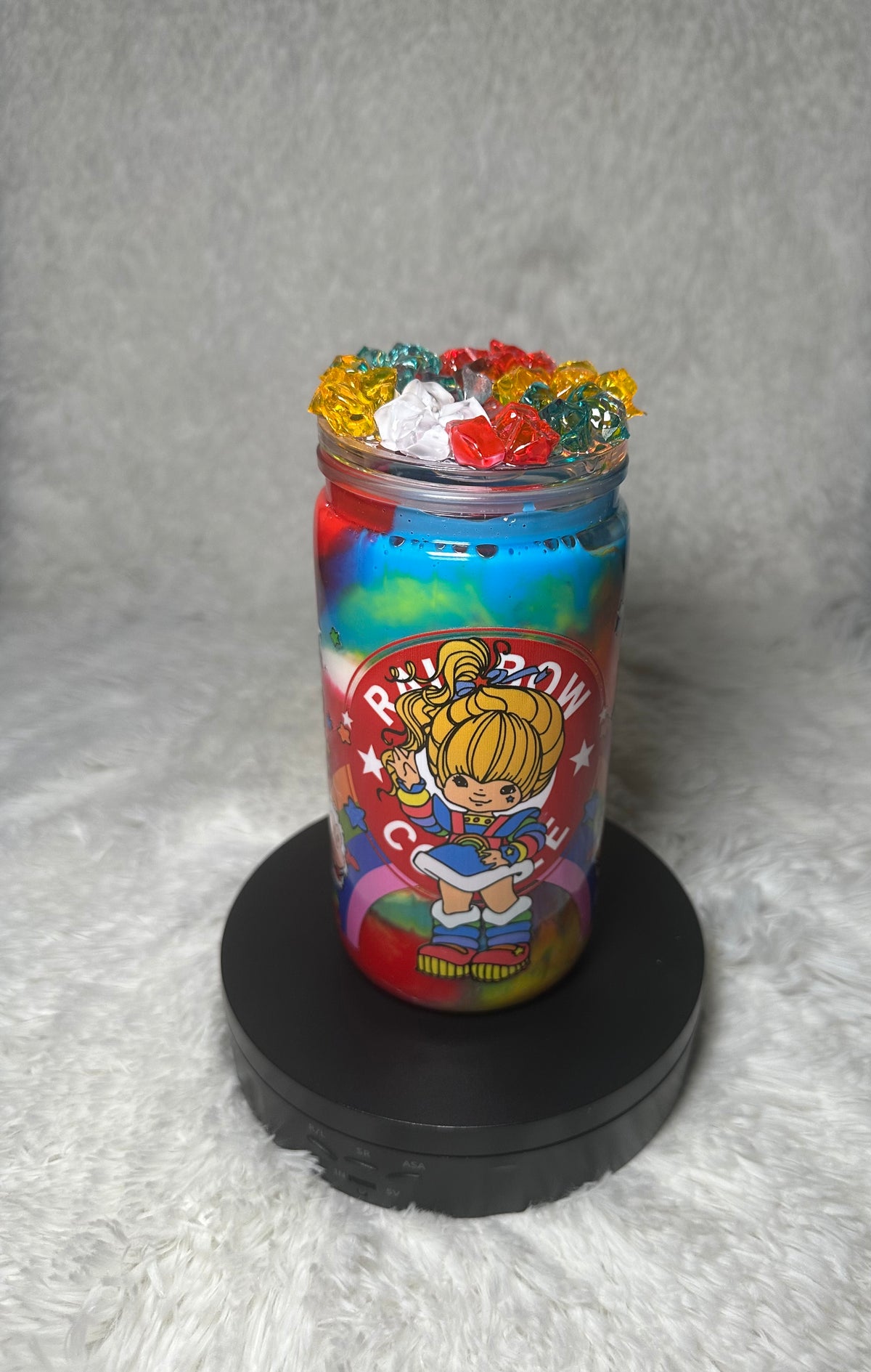 A colorful rainbow-themed jar filled with layered rainbow-colored material and topped with small multicolored flower-like decorations, displayed on a black round base against a soft gray background.