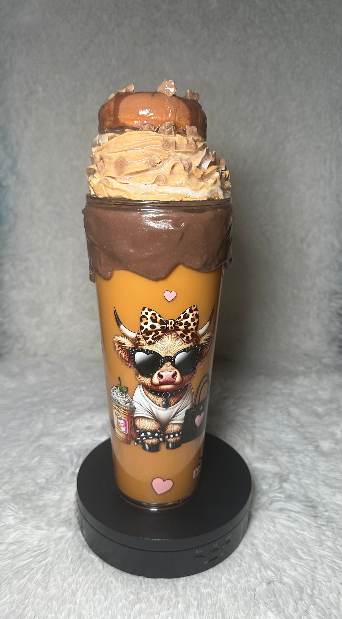 A custom-printed clear plastic tumbler cup with a cartoon cow character wearing sunglasses and a leopard-print bow tie, filled with a caramel-colored drink and topped with whipped cream and a donut decoration, displayed on a black circular stand against a gray background.