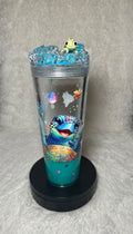 A clear plastic tumbler with a black base, decorated with a colorful cartoon sea turtle design and small ocean-themed graphics. The tumbler is filled with blue liquid and topped with a small green frog figurine, set against a gray textured background.