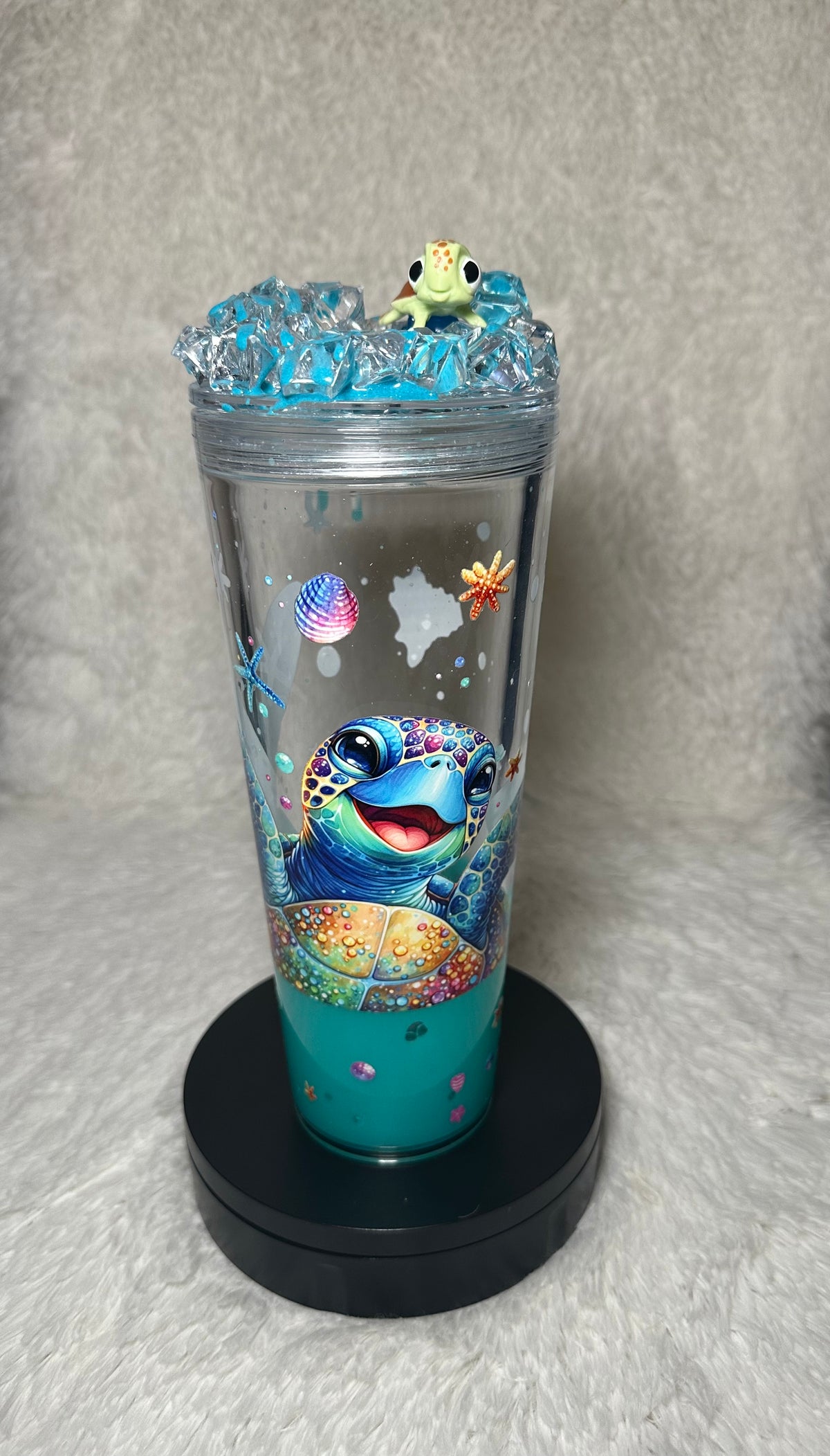 A clear plastic tumbler with a black base, decorated with a colorful cartoon sea turtle design and small ocean-themed graphics. The tumbler is filled with blue liquid and topped with a small green frog figurine, set against a gray textured background.