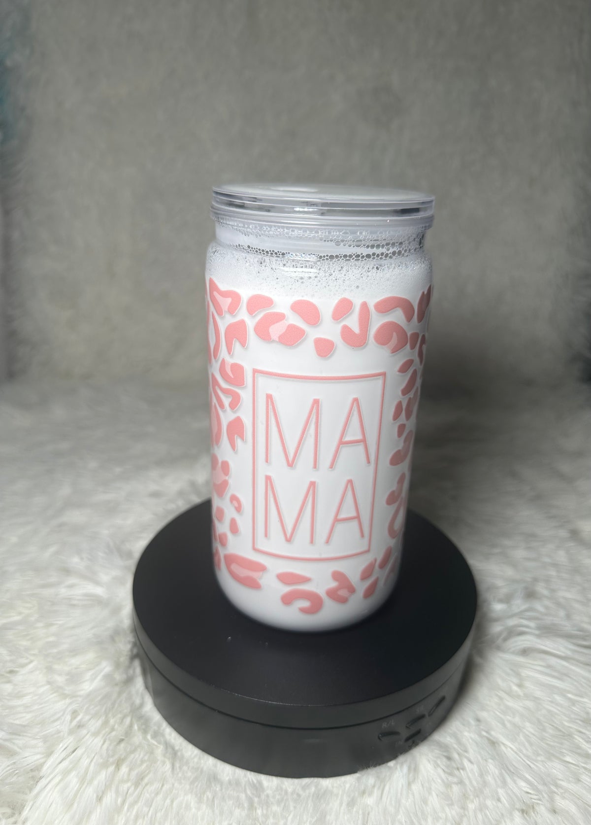 A white insulated tumbler with a clear lid, decorated with a pink leopard print and the word 'MAMA' in a pink square frame, displayed on a black circular base against a soft gray background.