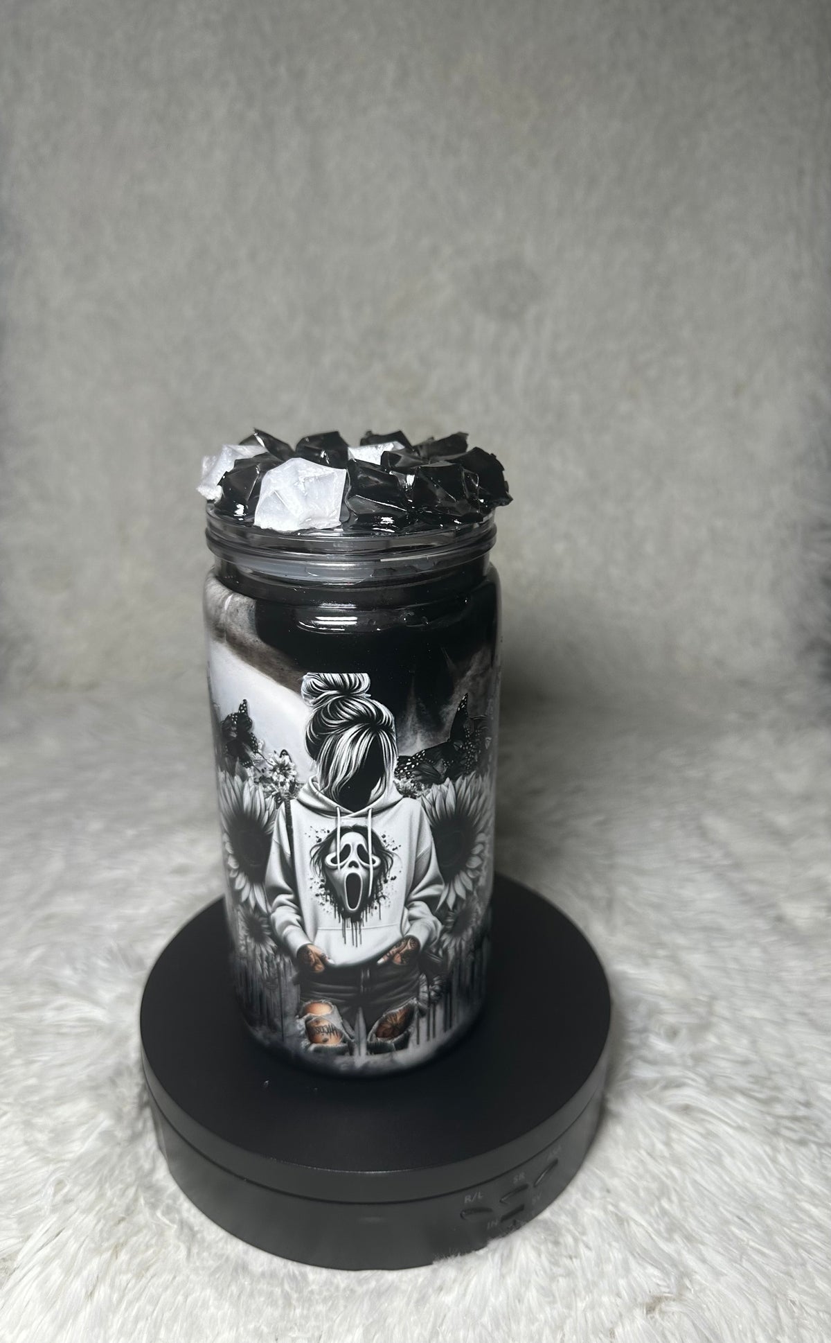A clear glass jar filled with black wax and topped with black wax flakes, featuring a detailed black and white graphic of a screaming face character, displayed on a black circular base against a gray textured background.