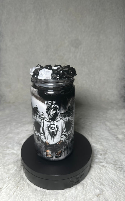 A clear glass jar filled with black wax and topped with black wax flakes, featuring a detailed black and white graphic of a screaming face character, displayed on a black circular base against a gray textured background.