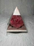 A pyramid-shaped resin keepsake or decorative object with a pink rose arrangement inside, set on a white faux fur surface.