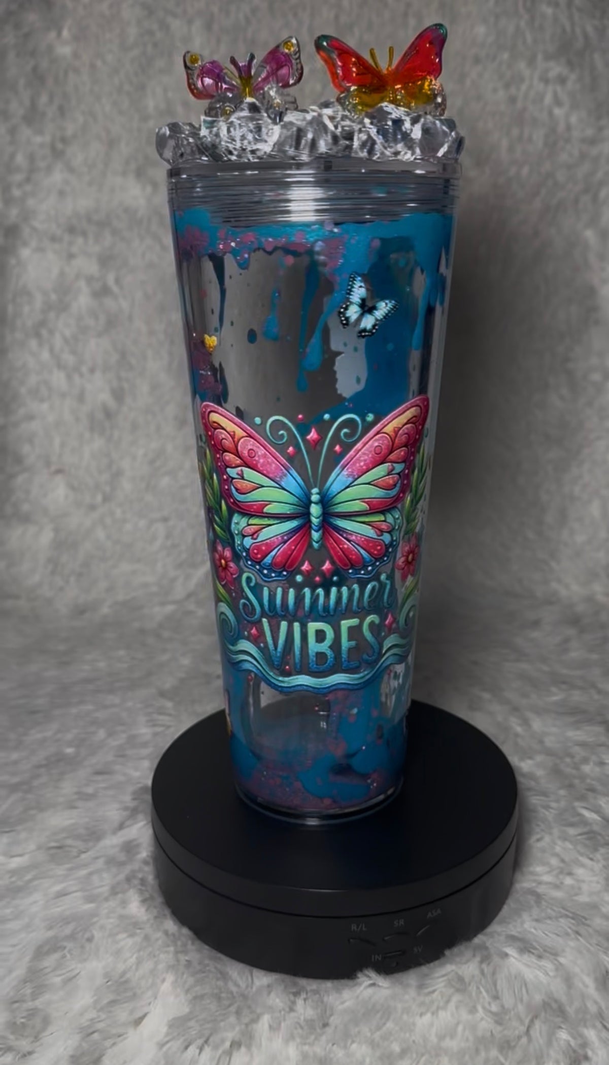 A clear insulated tumbler with a printed blue and pink butterfly design and the words 'Summer VIBES' on the front, topped with a clear lid and small decorative butterflies, displayed on a black circular stand against a gray textured background.